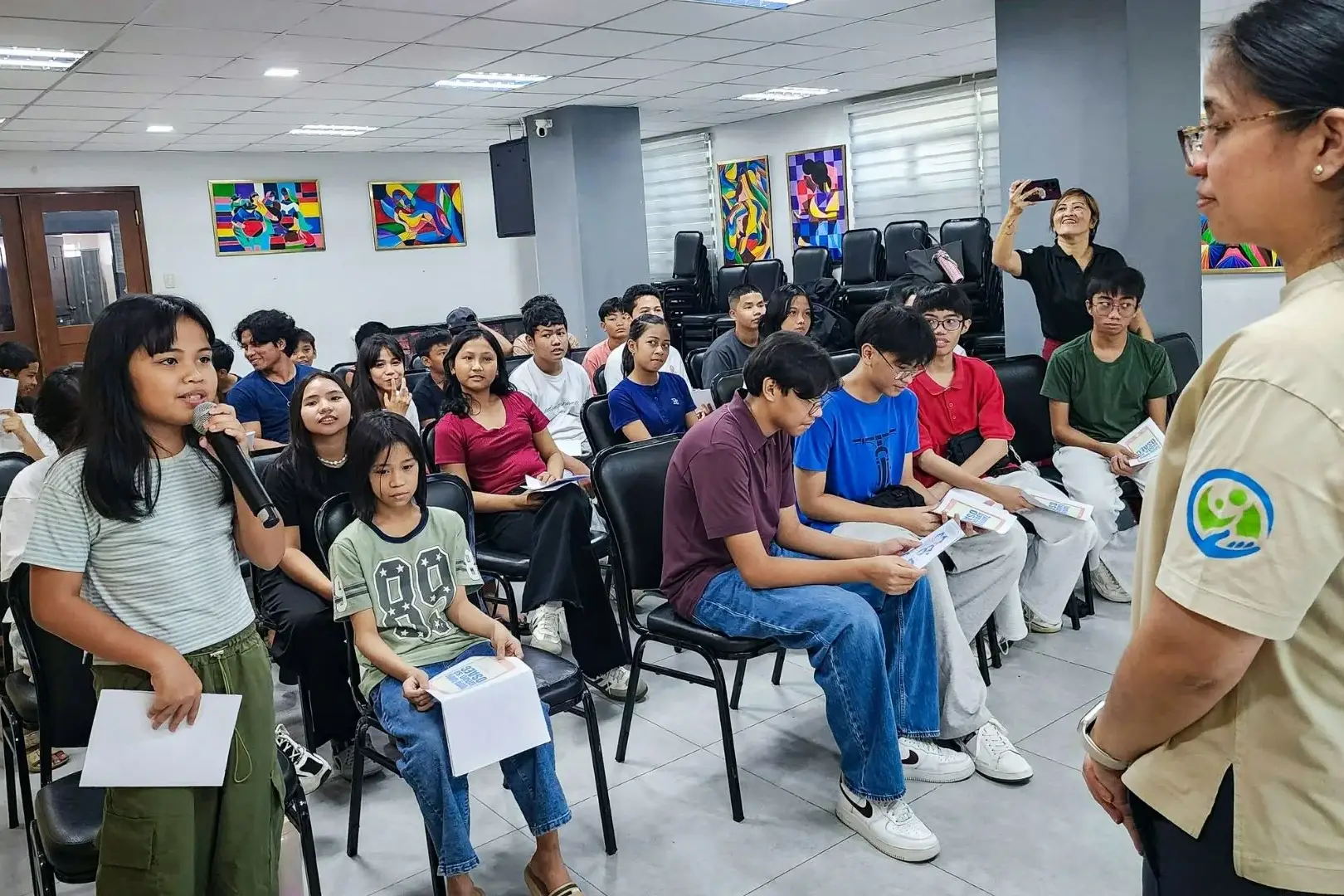 A young girl speaks into a microphone in a classroom, holding a paper. Students sit attentively, some holding papers. A woman in watches.