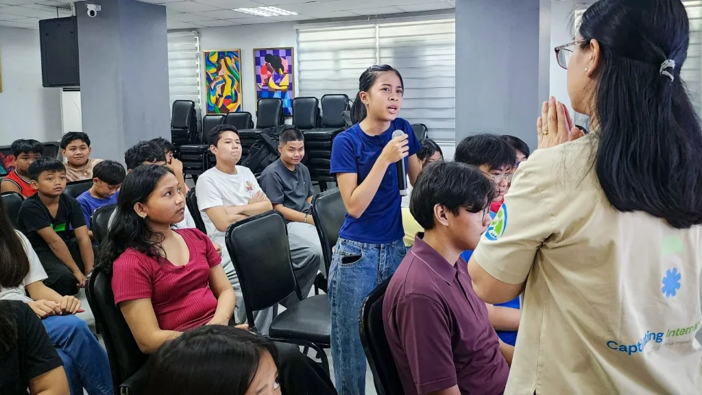 A young woman in a blue shirt stands, holding a microphone, speaking to a seated group in a classroom. Participants appear engaged and attentive.