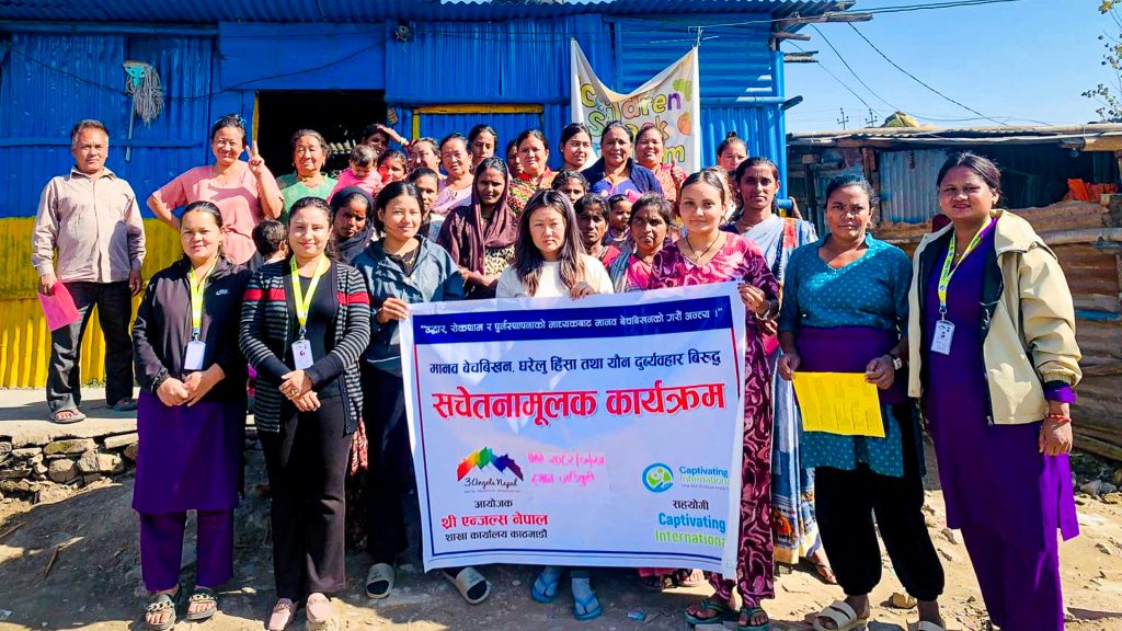A group of people, mostly women, pose outside a blue structure holding a banner with Nepali text and logos for an awareness event.