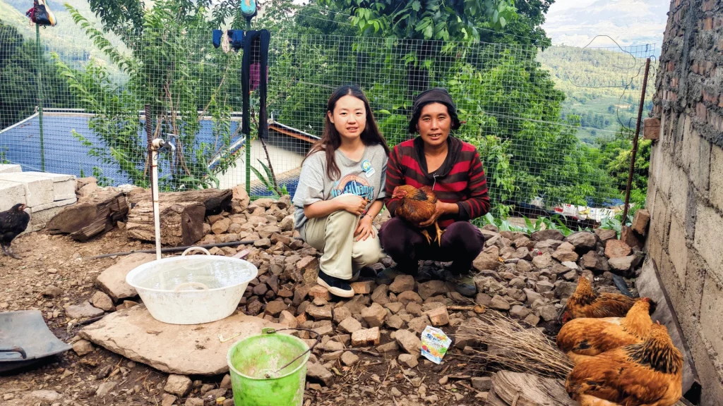 Two women sitting on the ground, engaged in conversation, surrounded by grass and a few scattered flowers.