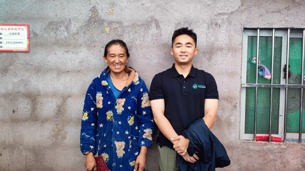 A man and a woman stand side by side against a textured wall, both smiling and dressed casually.