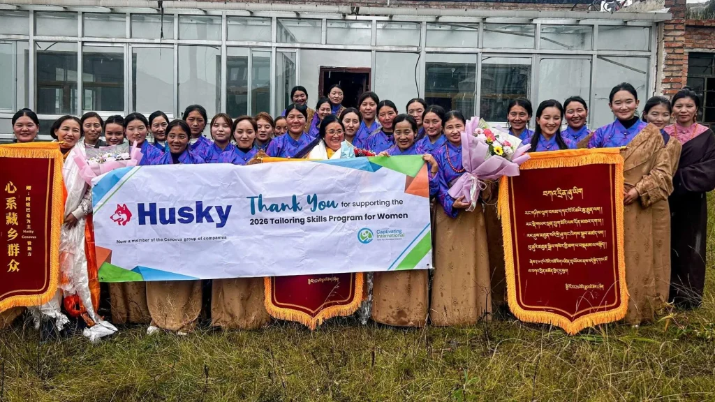 Smiling women pose outdoors with a “Thank You” banner and red plaques, celebrating a tailoring skills program for women in rural China.