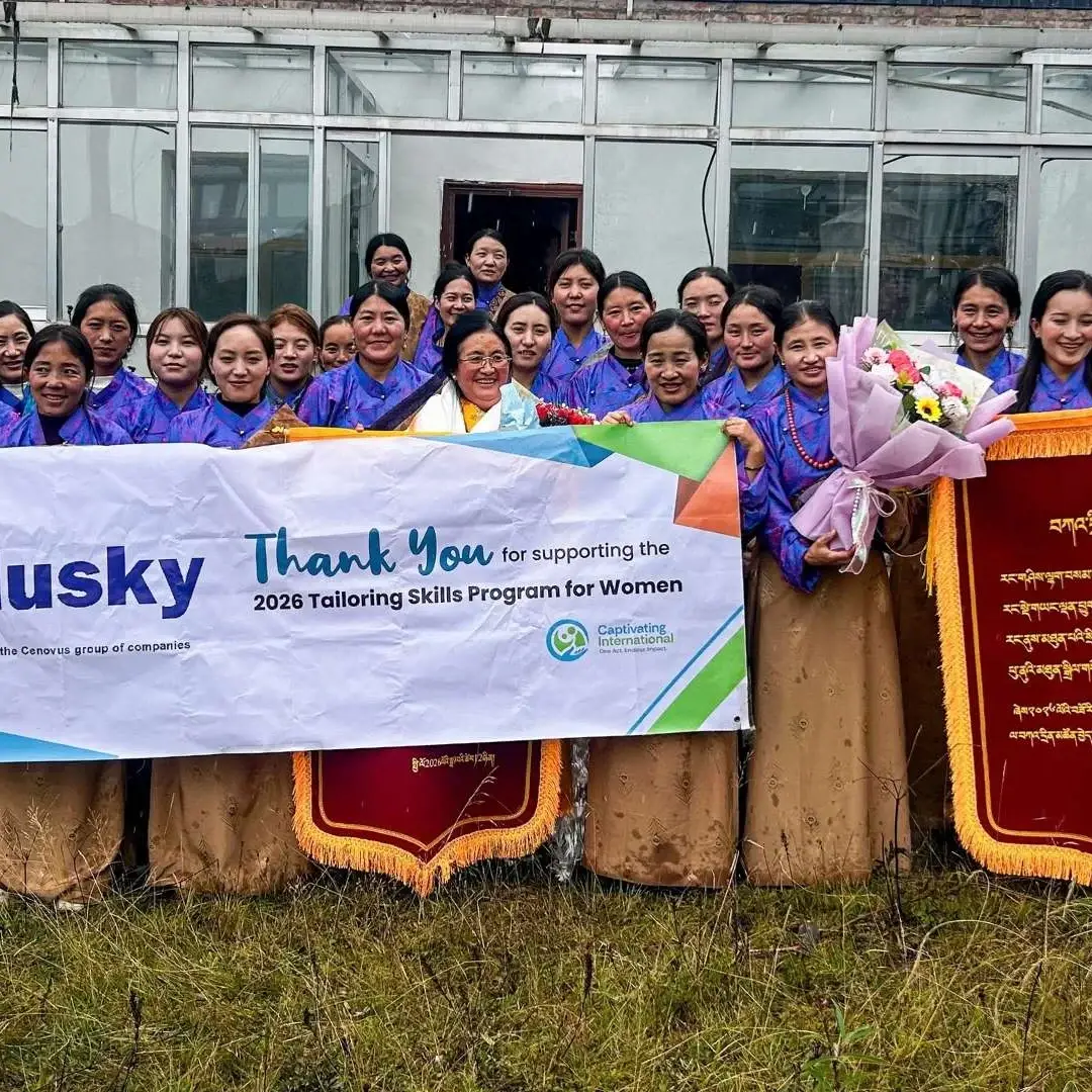 Smiling women pose outdoors with a “Thank You” banner and red plaques, celebrating a tailoring skills program for women in rural China.