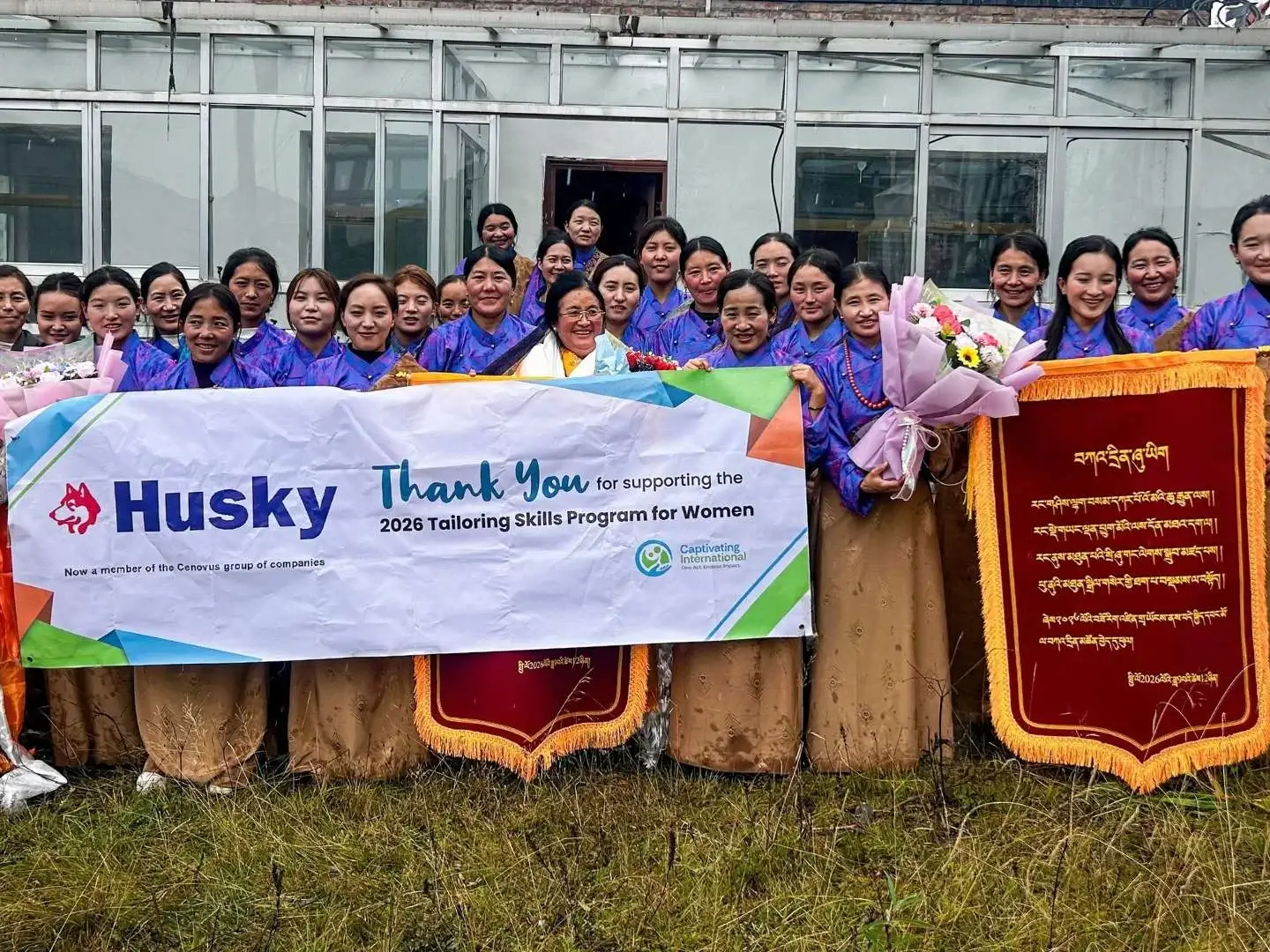 Smiling women pose outdoors with a “Thank You” banner and red plaques, celebrating a tailoring skills program for women in rural China.