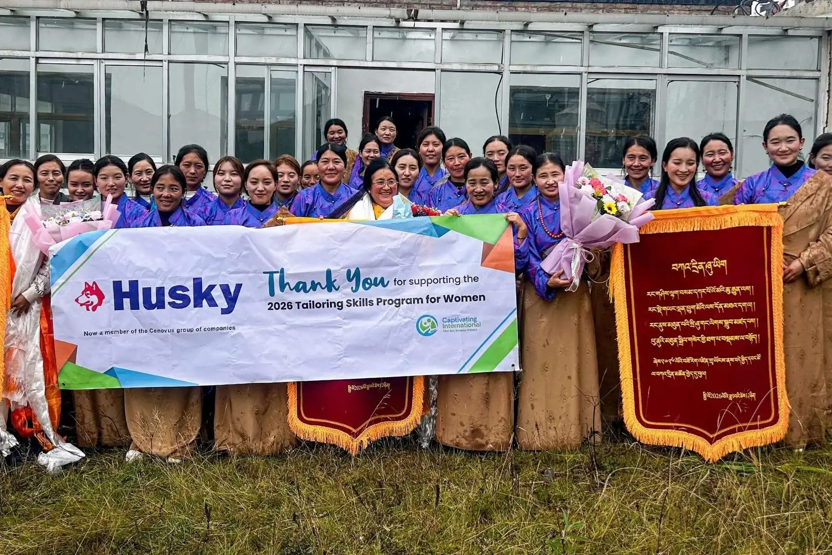 Smiling women pose outdoors with a “Thank You” banner and red plaques, celebrating a tailoring skills program for women in rural China.