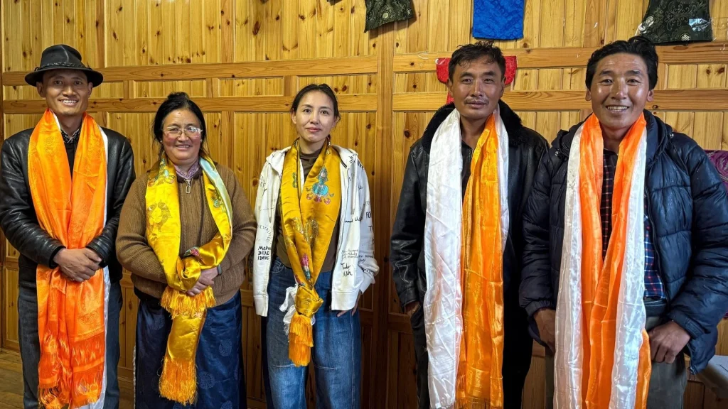 Five smiling adults in rural China pose in a wood-paneled room, wearing colorful ceremonial scarves in orange, yellow, and white; the group portrait feels warm and welcoming.