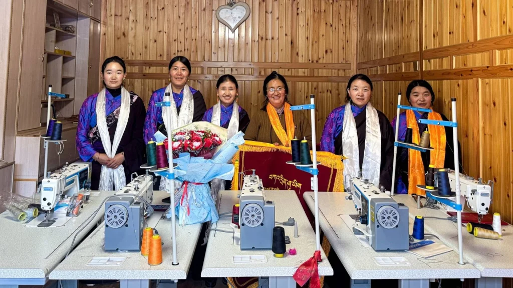 Six Chinese women in colorful traditional clothing pose behind sewing machines in a wood-paneled workshop, beside a bouquet of flowers, creating a warm, celebratory mood.