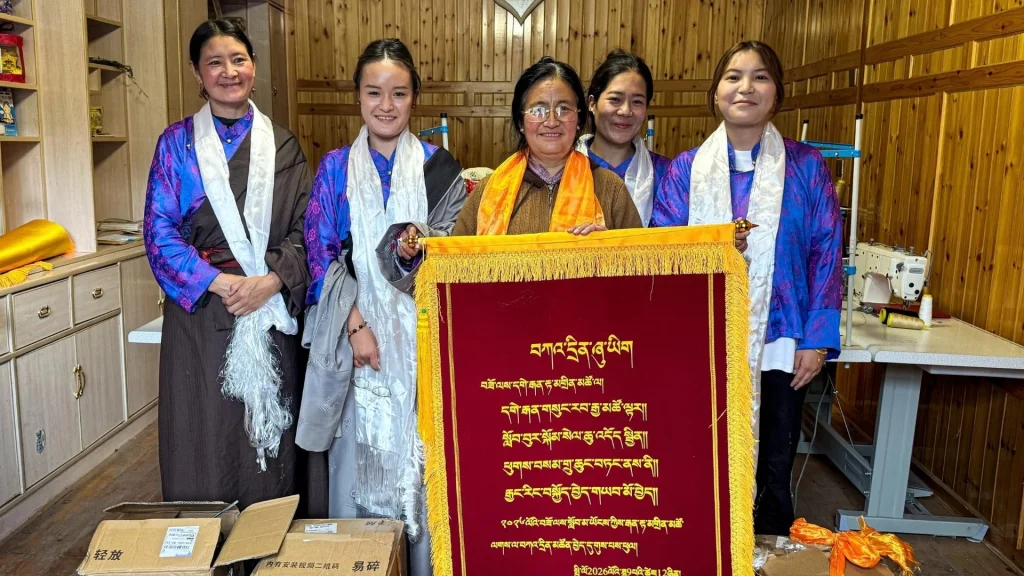 Five Chinese women in colorful traditional dress pose behind a red, gold-fringed banner with ornate script in a wood-paneled room, conveying pride and celebration.