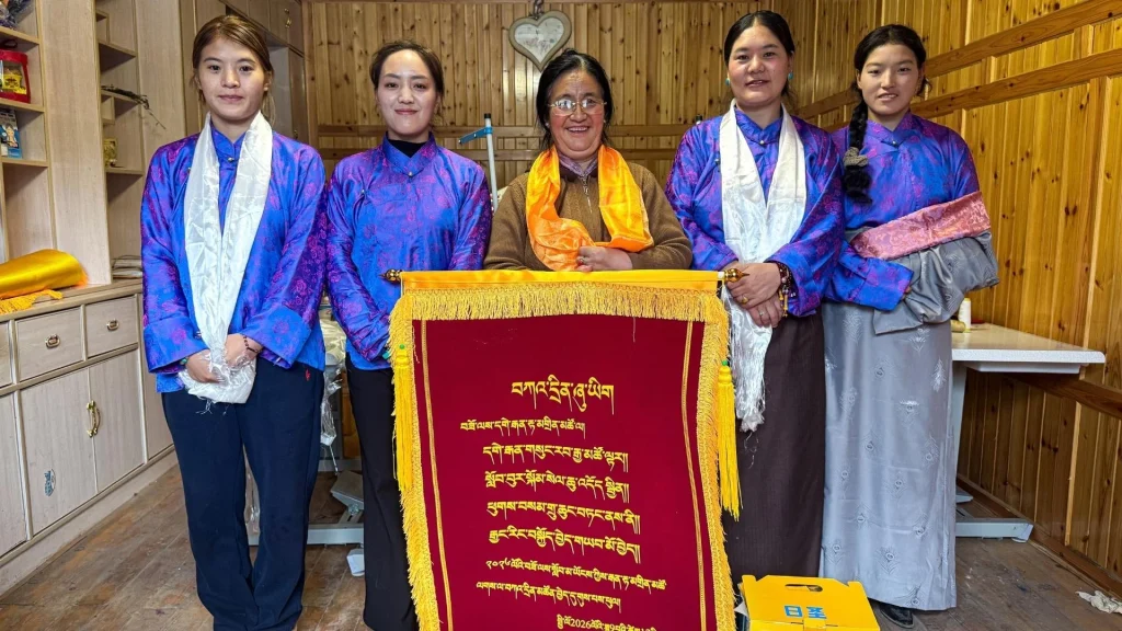 Five women in blue and orange traditional clothing pose behind an ornate red-and-gold Tibetan-style ceremonial banner in a warm wooden room, smiling proudly.