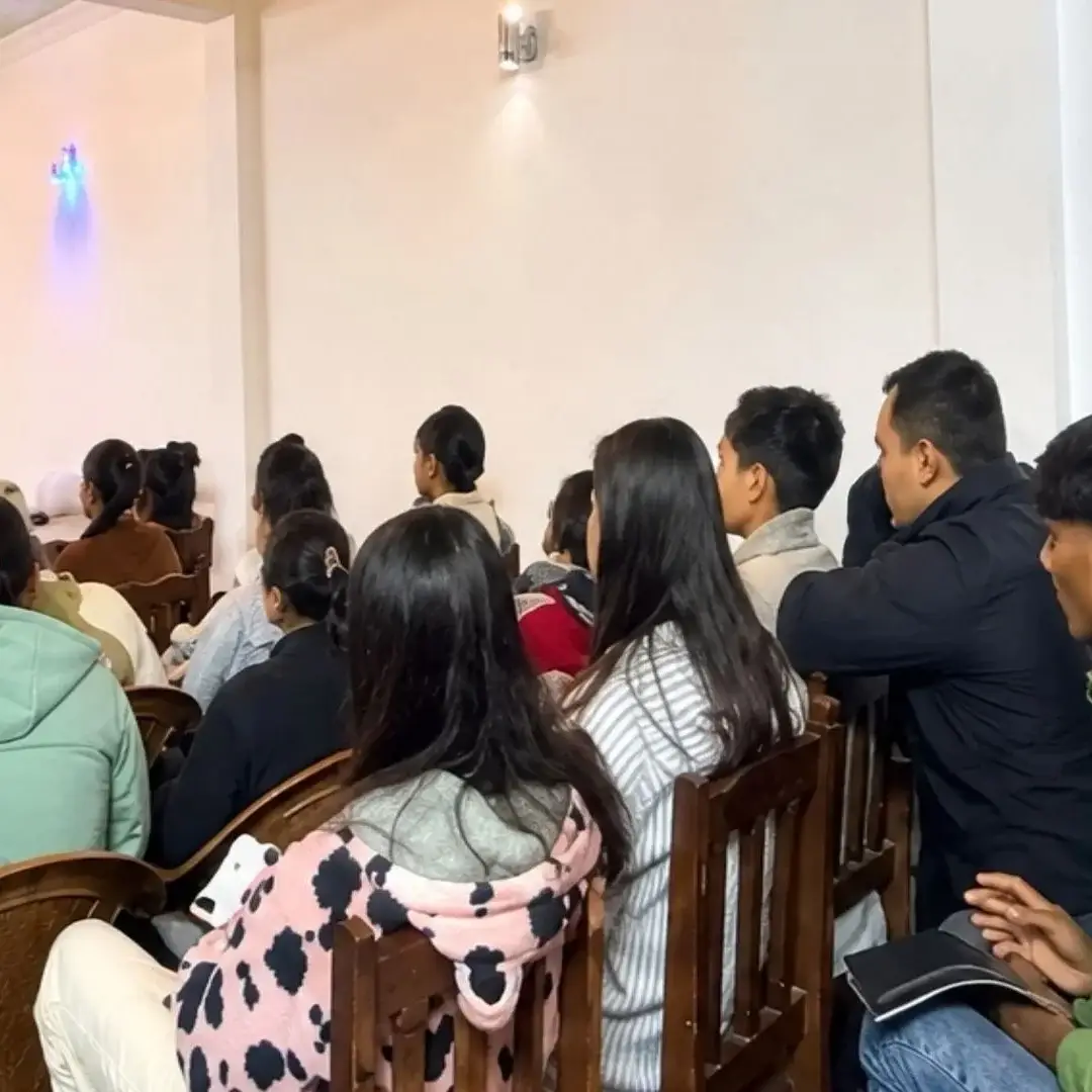 A woman stands at the front of a room giving a presentation to a group of students sitting in wooden chairs, all facing toward her in a bright hall in Nepal.