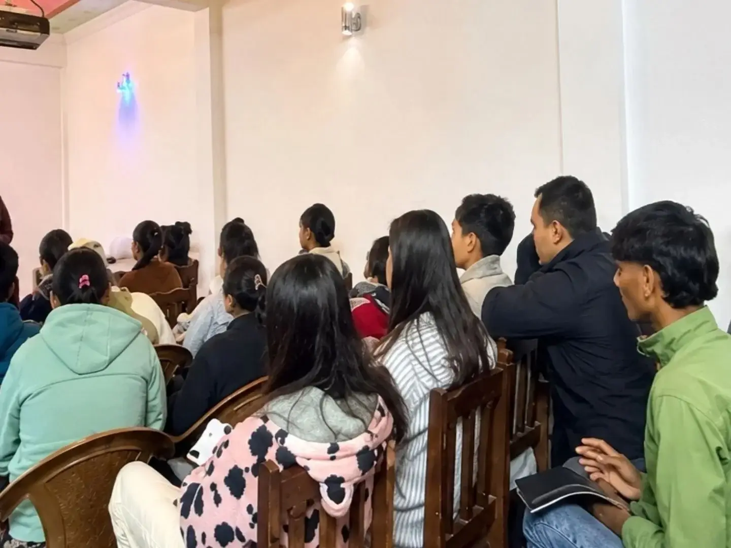 A woman stands at the front of a room giving a presentation to a group of students sitting in wooden chairs, all facing toward her in a bright hall in Nepal.