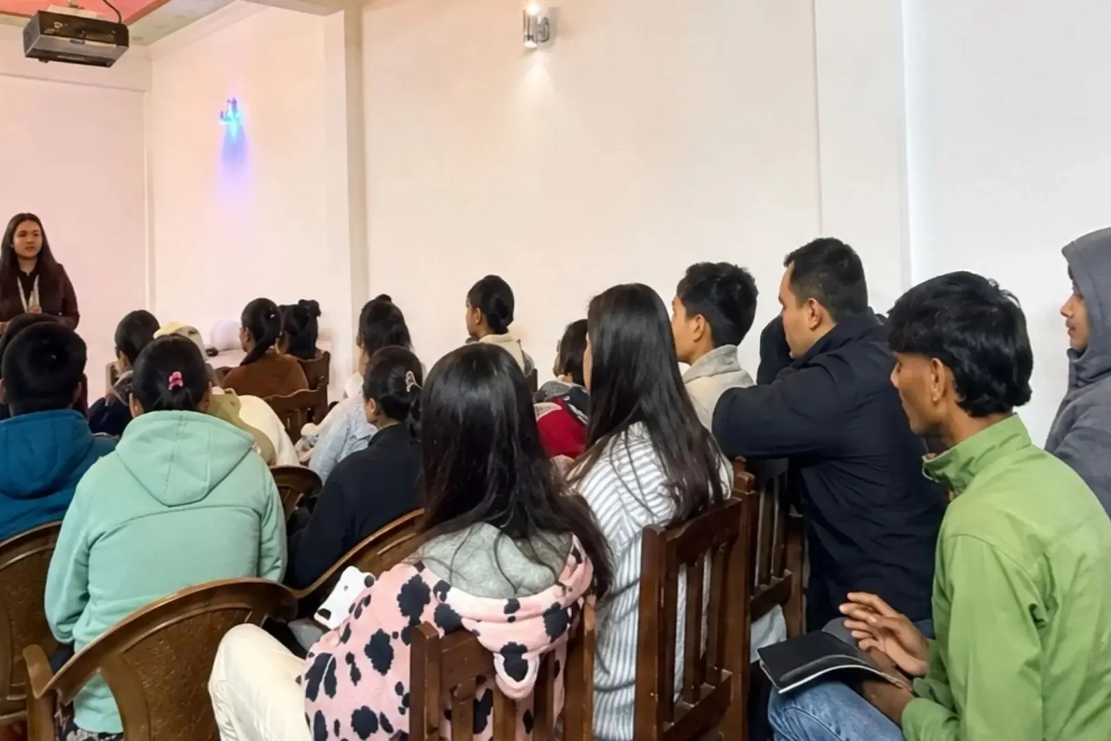 A woman stands at the front of a room giving a presentation to a group of students sitting in wooden chairs, all facing toward her in a bright hall in Nepal.