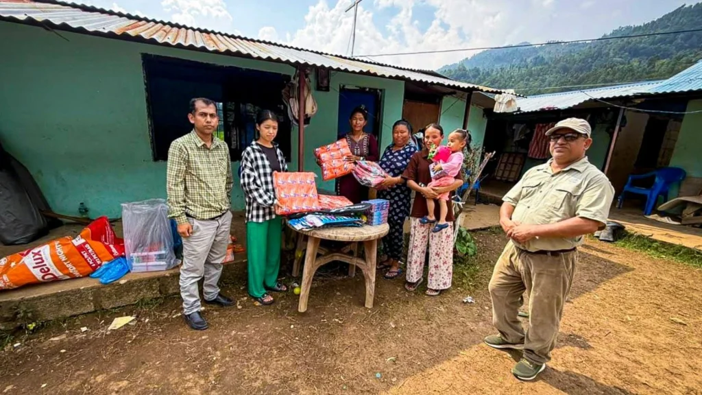 A group of adults and a child stand outside a house in a rural area, with some people holding packages of goods placed on a small wooden table.