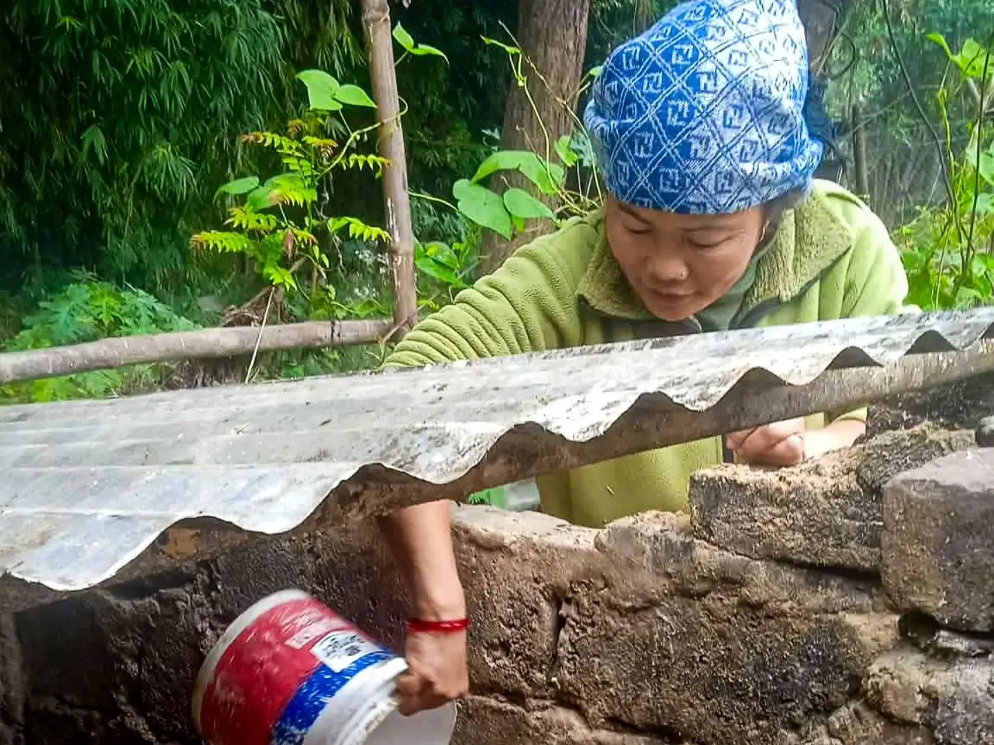 A woman in a blue patterned headscarf and green fleece pours liquid from a plastic bucket into a stone pigpen containing a brown pig in a rural area.