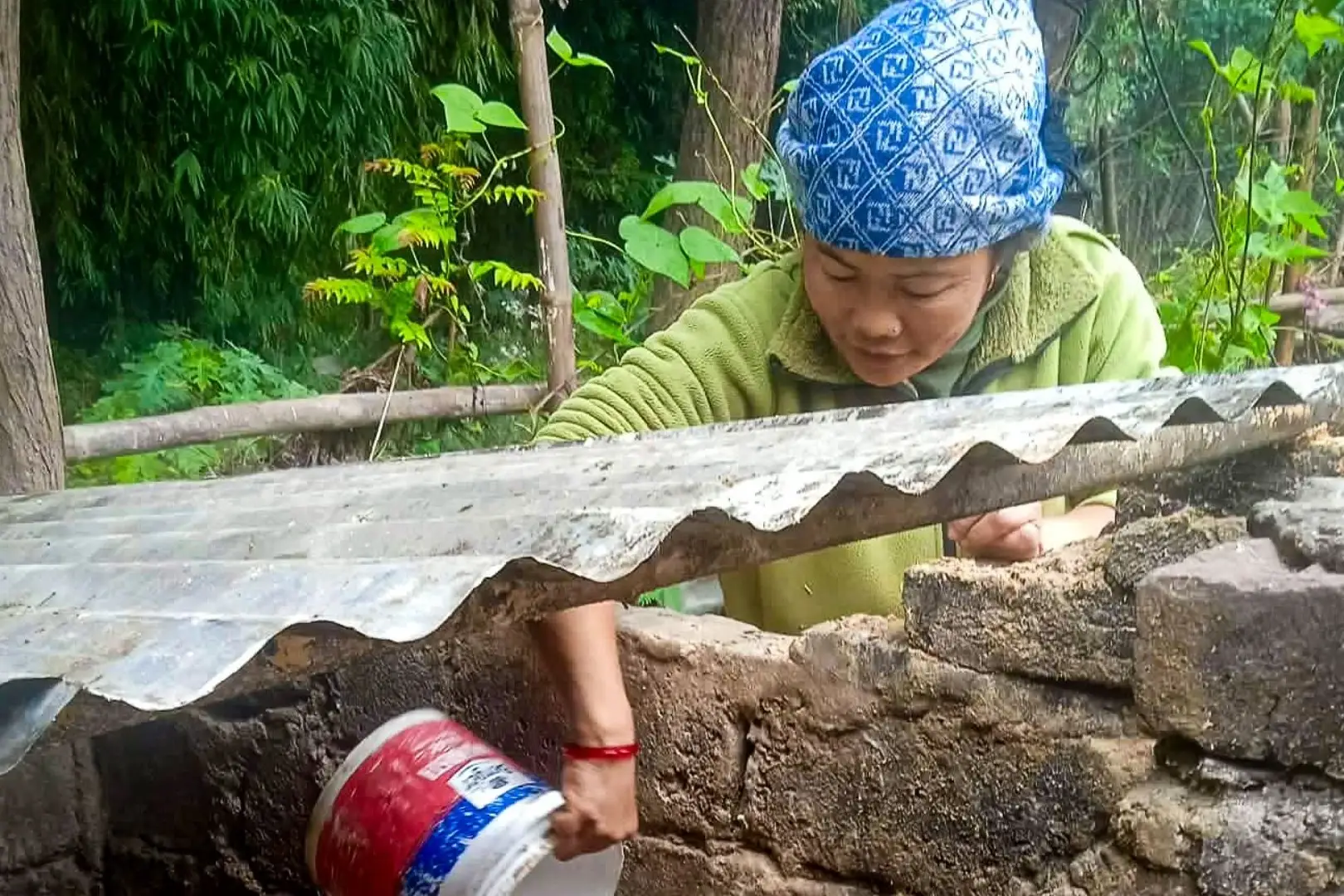 A woman in a blue patterned headscarf and green fleece pours liquid from a plastic bucket into a stone pigpen containing a brown pig in a rural area.
