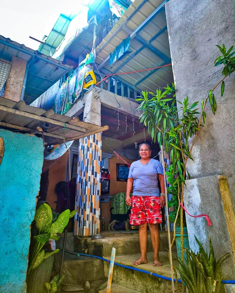 A woman poses in front of a house with a sign, showcasing a welcoming expression and a friendly demeanor.