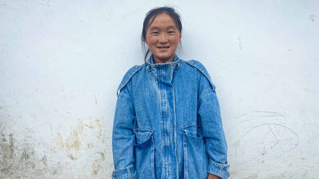 A woman in a denim jacket stands against a plain wall, smiling softly.