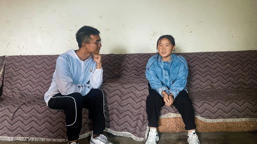 A man and woman are seated on a couch, engaged in conversation, with a wall behind them in a casual living space.