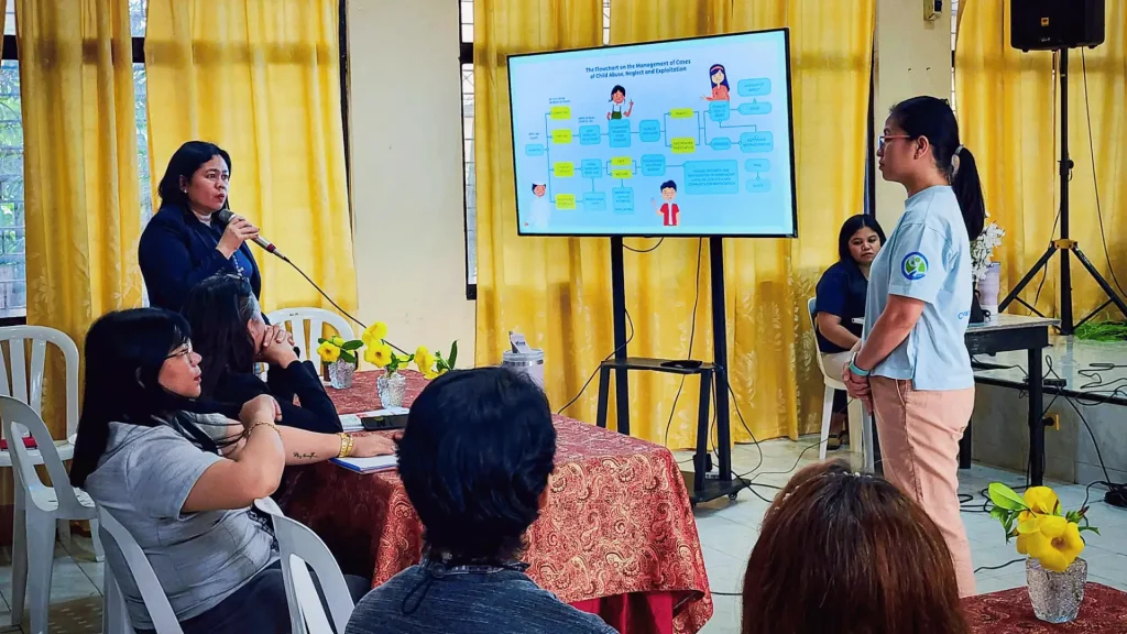 People sitting at tables, engaged in a discussion while a screen on the wall shows a presentation.
