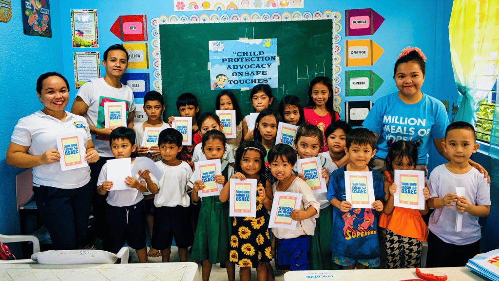 A group of smiling children and facilitators proudly holding their books in front of a colorful classroom.