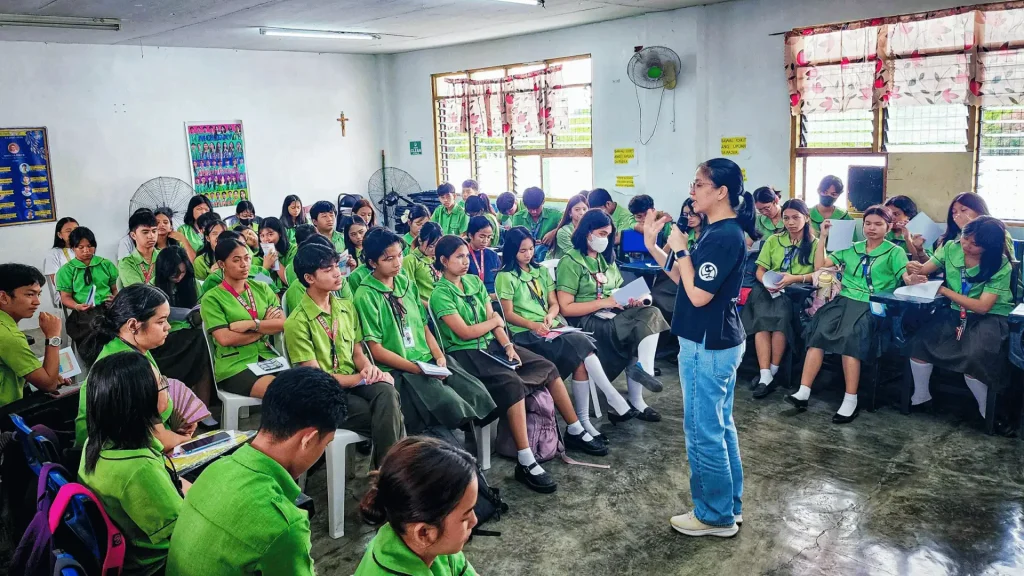A classroom scene featuring students in green shirts, sitting at desks and participating in a group discussion.