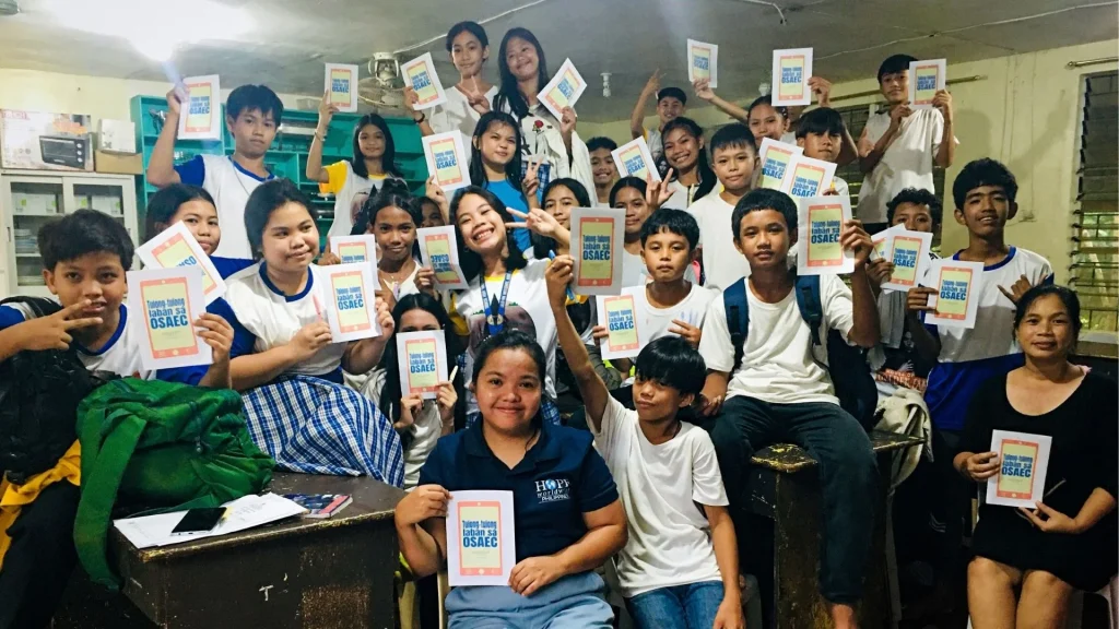 A large group of students and a teacher in a classroom holding up identical flyers that read Tulong-tulong laban sa OSAEC, smiling at the camera.