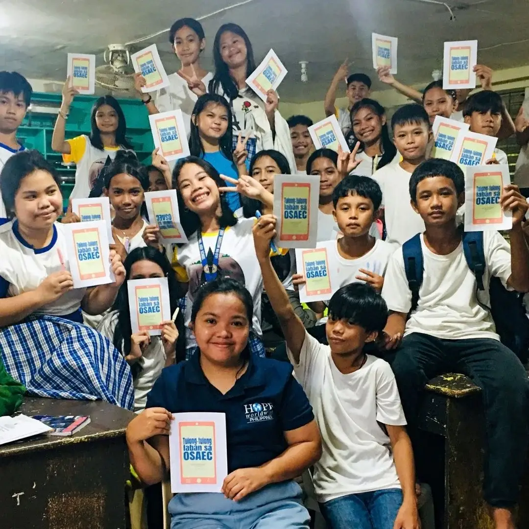 A large group of students and a teacher in a classroom holding up identical flyers that read Tulong-tulong laban sa OSAEC, smiling at the camera.