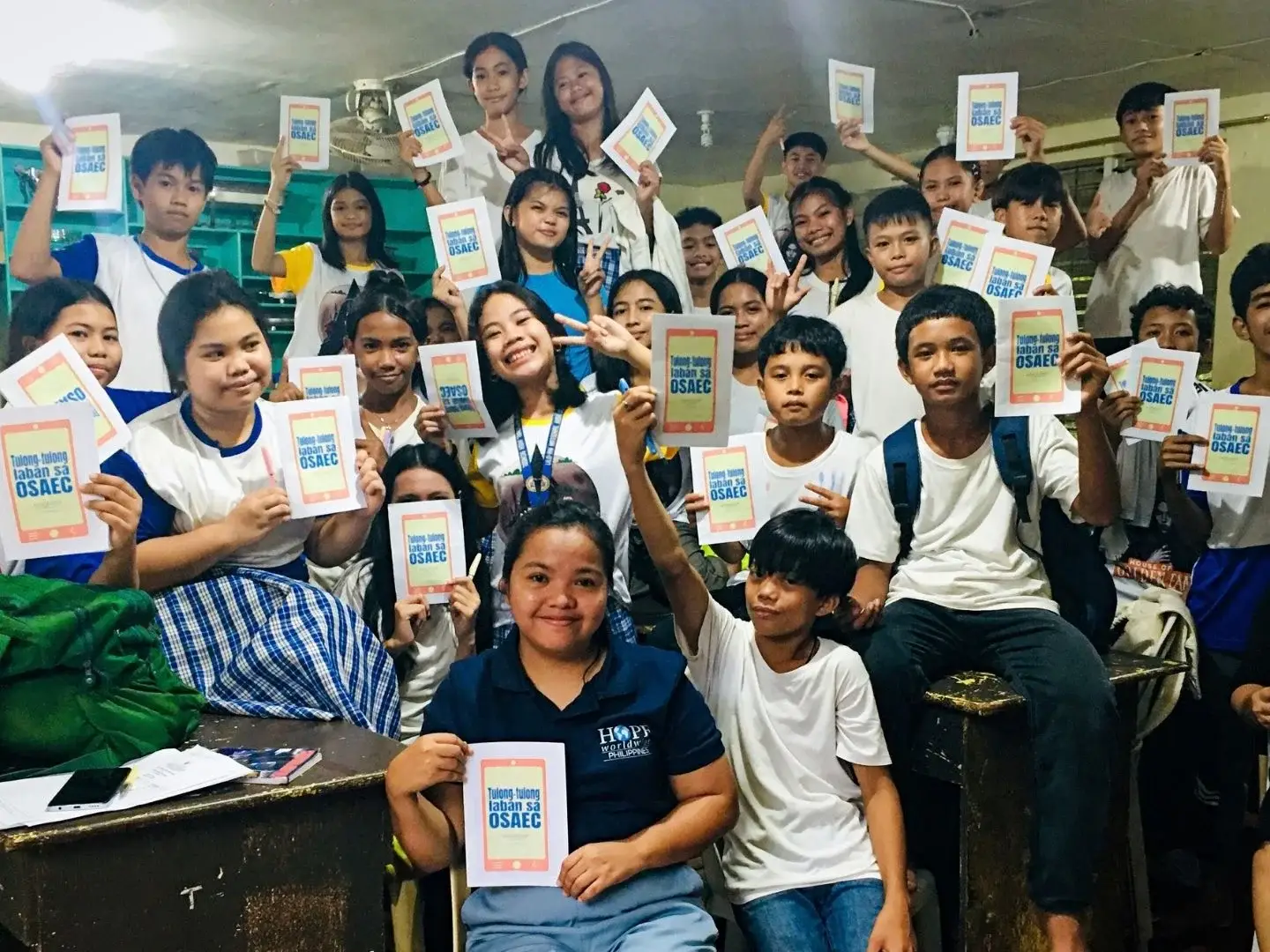 A large group of students and a teacher in a classroom holding up identical flyers that read Tulong-tulong laban sa OSAEC, smiling at the camera.