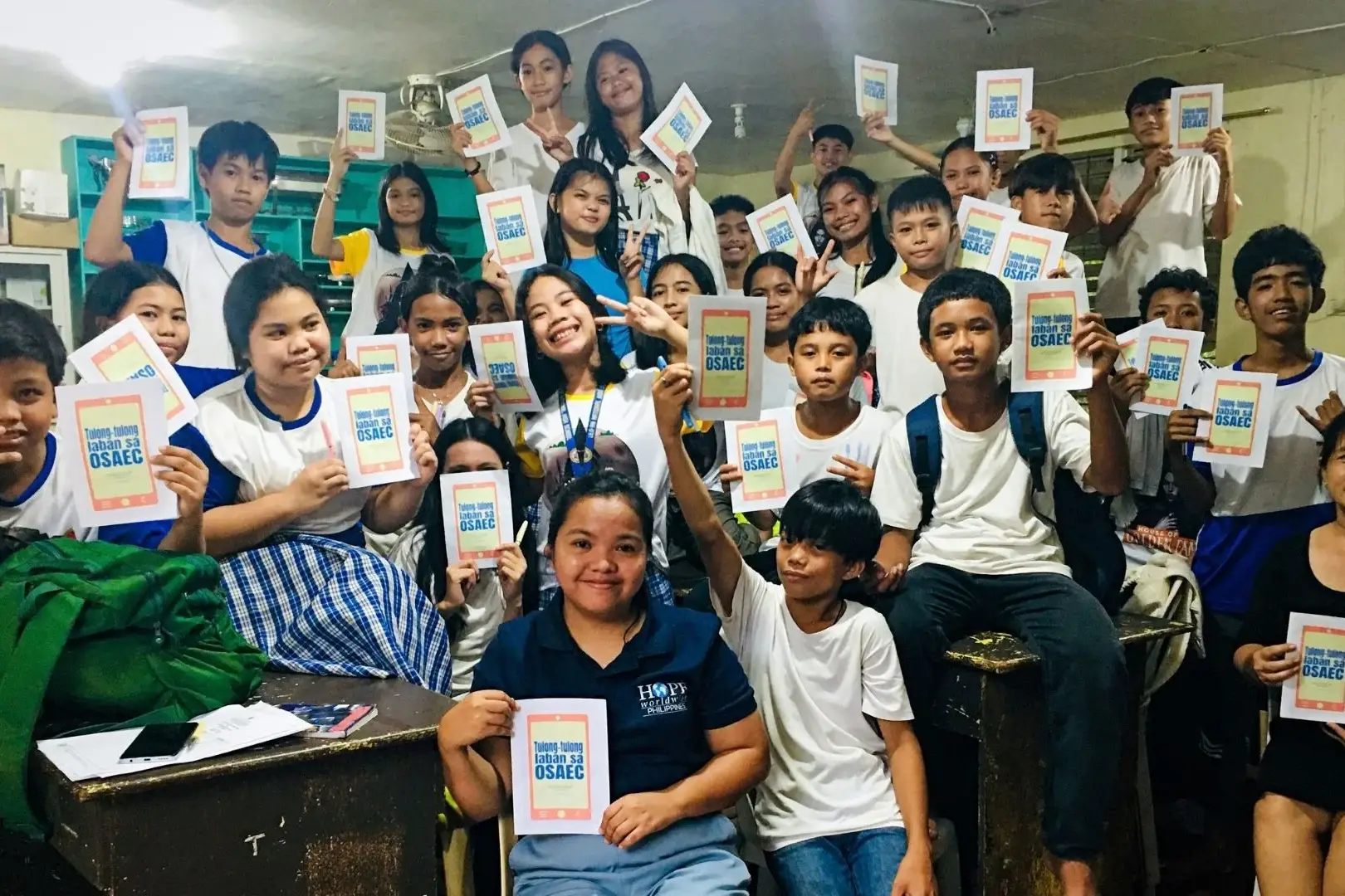 A large group of students and a teacher in a classroom holding up identical flyers that read Tulong-tulong laban sa OSAEC, smiling at the camera.