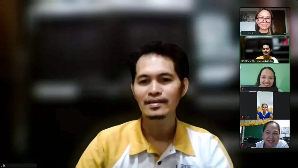 A man in a yellow and white collared shirt speaks during a video conference with four other participants visible in a sidebar on the right side.
