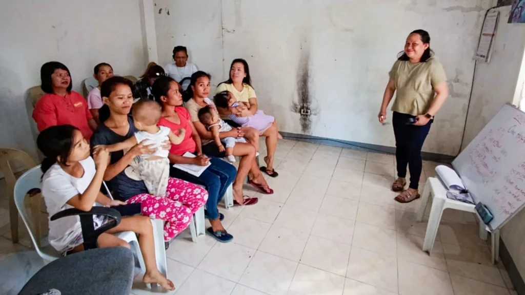 A woman stands before a group of seated women and children in a room, presenting information on a whiteboard during a community health meeting.