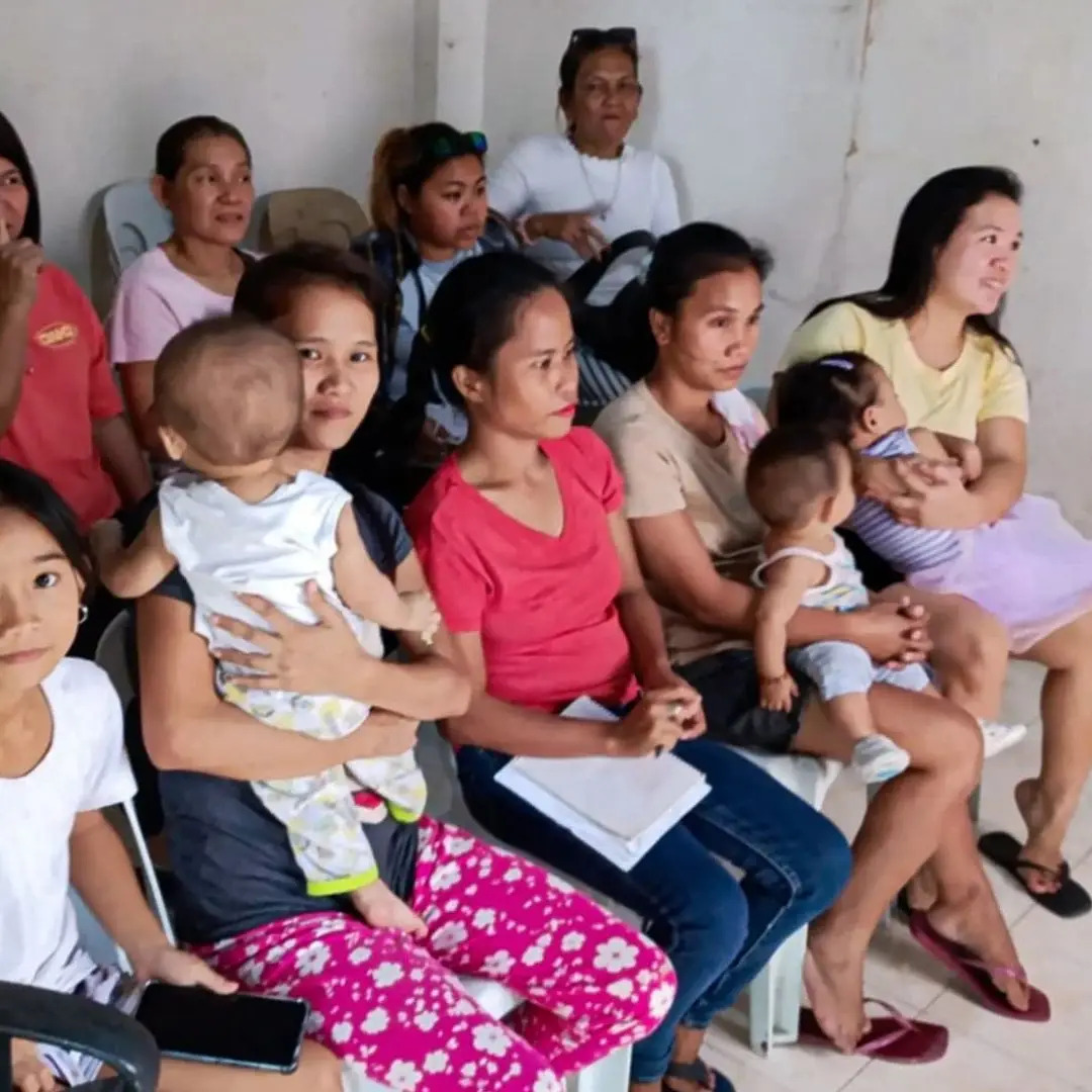 A group of women and children sitting on plastic chairs in a simple room, with some women holding infants and others looking toward the camera.