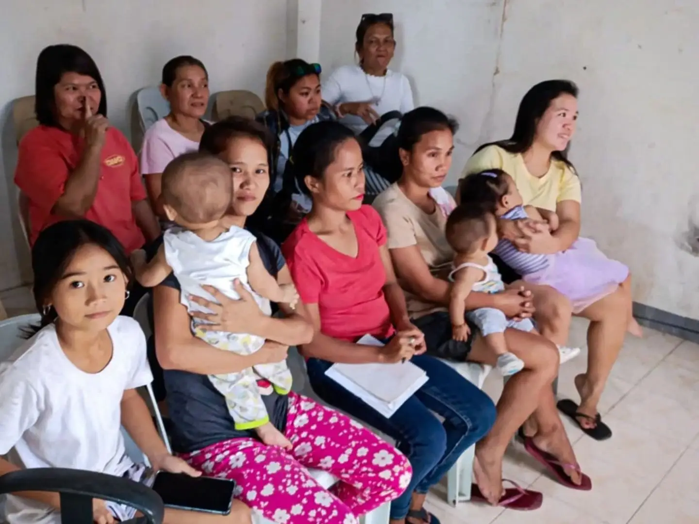A group of women and children sitting on plastic chairs in a simple room, with some women holding infants and others looking toward the camera.