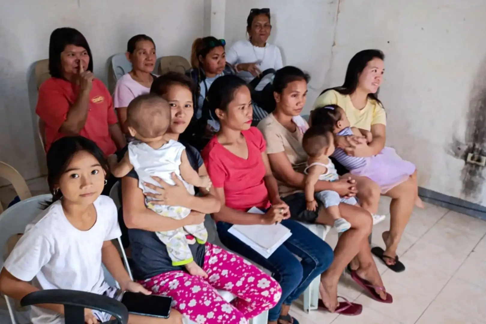 A group of women and children sitting on plastic chairs in a simple room, with some women holding infants and others looking toward the camera.