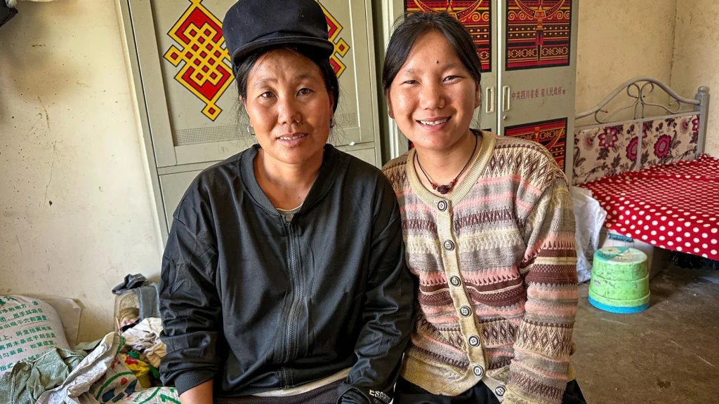Two women sit together indoors; one wears a black jacket and cap, while the other wears a patterned, button-up cardigan and a simple necklace.