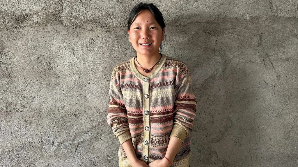 A young woman with dark hair smiles while wearing a patterned, button-up knit cardigan against a textured, grey unfinished concrete wall background.