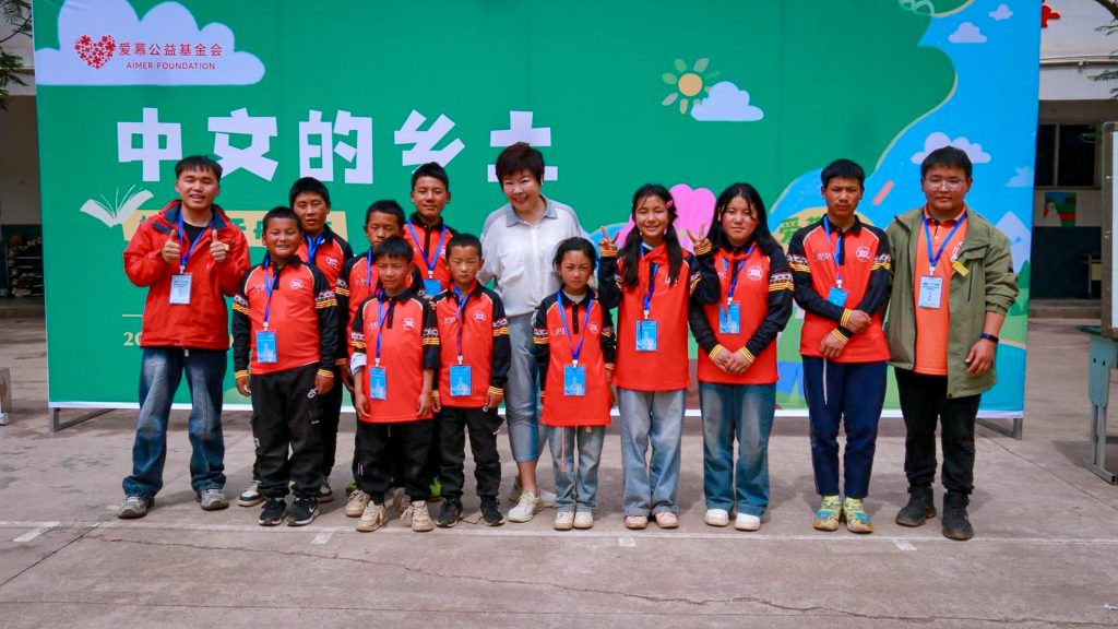A group of children and adults wearing matching red jackets and blue lanyards pose in front of a green event backdrop with Chinese text and logos.