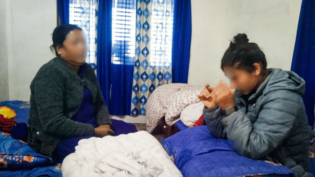 Two women on a bed in a softly lit room, smiling and talking.