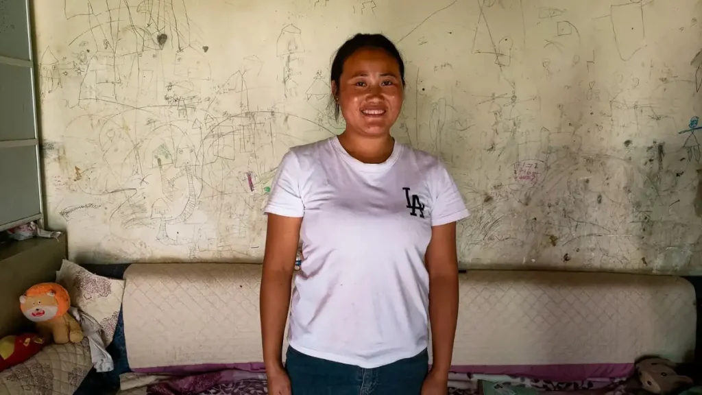 A woman wearing a white t-shirt stands in front of a neatly made bed.
