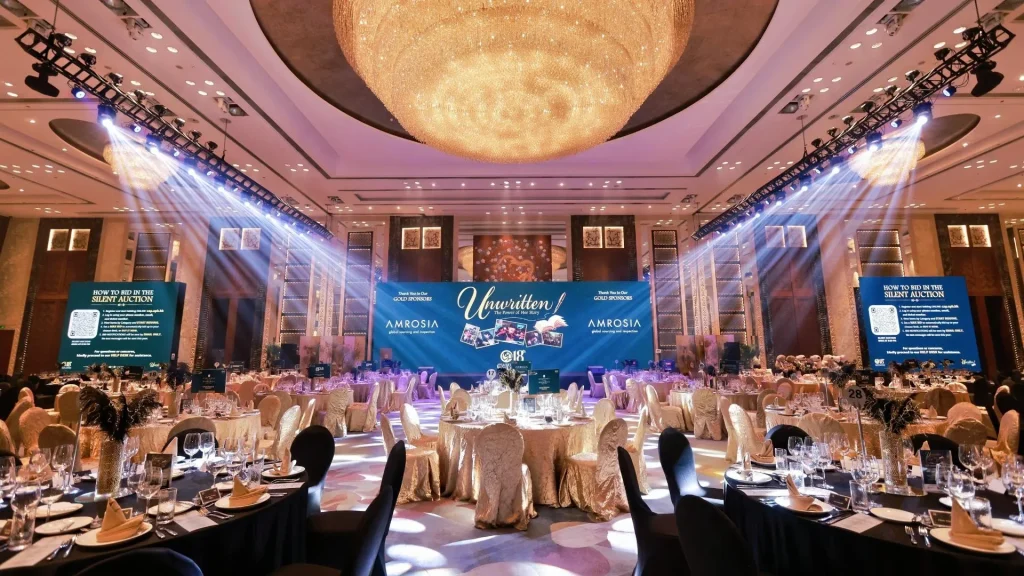 A lavish awards gala with guests seated at round tables beneath a glowing chandelier, while spotlights illuminate a blue Unwritten-themed stage.