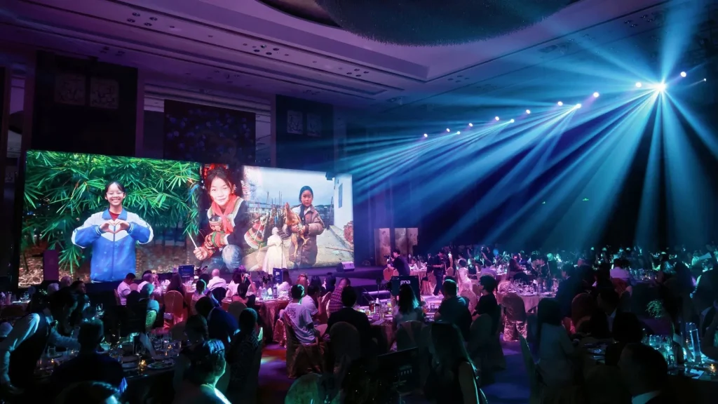 A packed audience watches performers in colorful traditional costumes on a large screen, while blue spotlights sweep across the lively indoor event.
