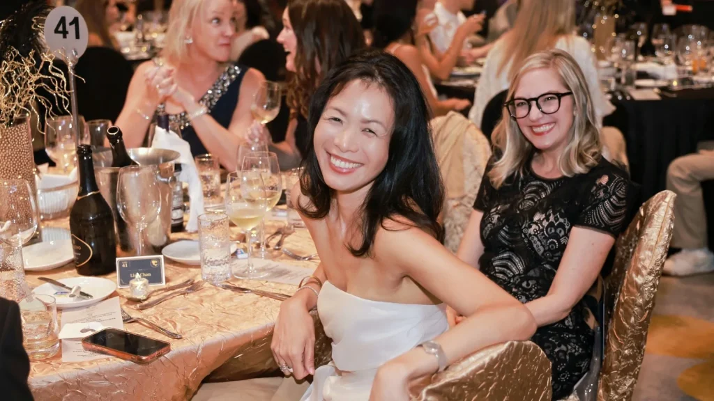 Two women smile at a lavish formal banquet, one in a white strapless dress and the other in black lace and glasses, amid warm, festive dinner décor.