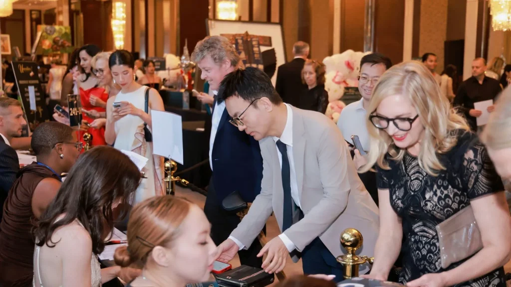 At a lively, warm-lit indoor event, guests gather around a display table as a man leans in and a smiling blonde woman examines items.