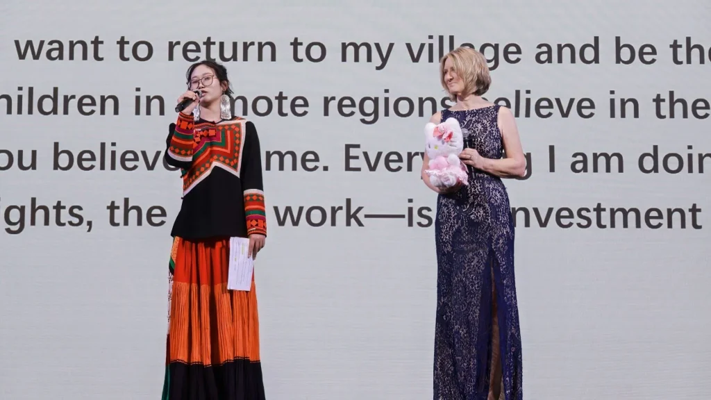 Two women stand onstage before a screen of large text. One speaks into a microphone in a vivid red-and-orange traditional outfit; the other holds a pink plush toy. The scene feels formal and hopeful.