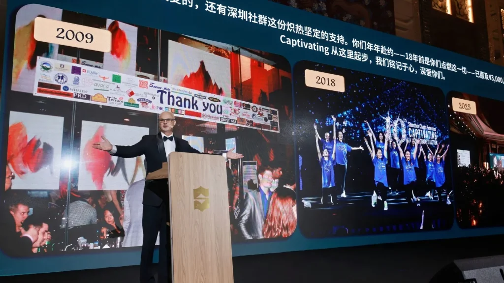 A man in a tuxedo speaks at a podium in front of a large screen displaying historical photos from 2009 and 2018 at a charity event in Shenzhen.