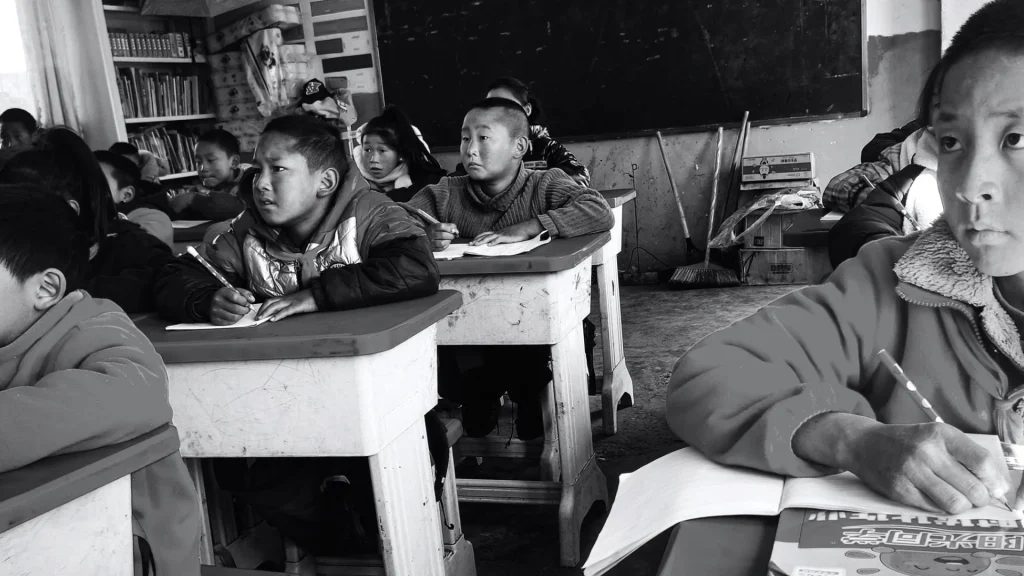Black-and-white classroom scene with children writing at worn desks, a chalkboard behind them, and a focused student in the foreground.