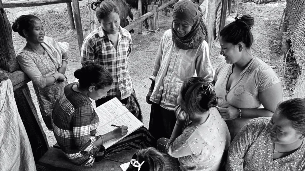 Black-and-white photo of women gathered outdoors, discussing paperwork as one woman writes; the candid scene conveys community and support.