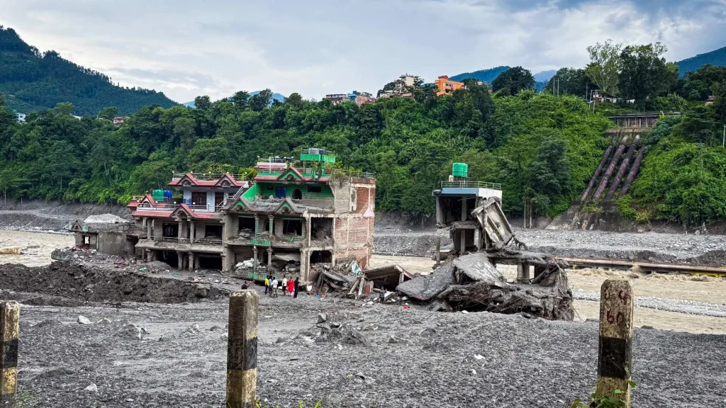 Severely damaged riverside buildings and concrete debris fill a gray riverbed, with tiny people, lush forested mountains, and cloudy skies behind in Nepal.
