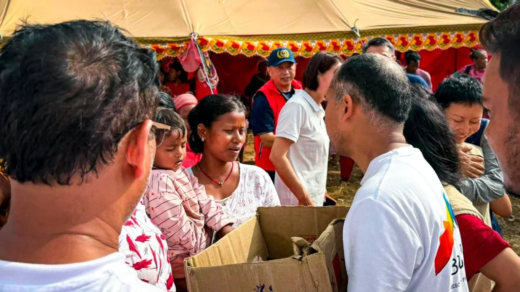 Crowded community event under a decorated tent, where volunteers distribute cardboard boxes to families, creating a busy, compassionate atmosphere in Nepal.