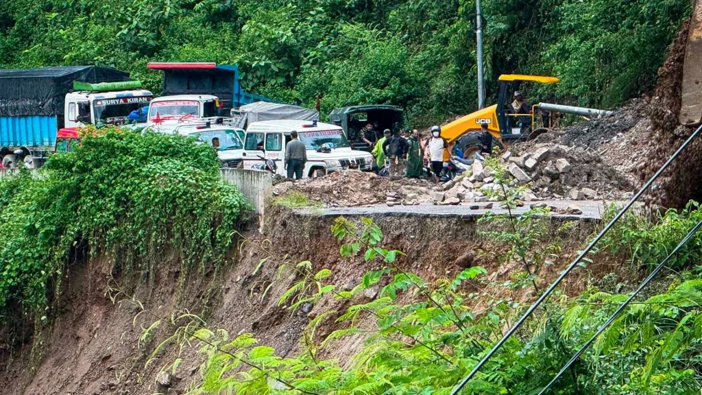 Roadside landslide cleanup with an excavator and workers clearing rubble beside vehicles, surrounded by lush tropical forest and steep, eroded slopes in Nepal.